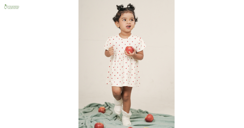 Baby girl in white organic cotton dress standing among red apples, representing GOTS-certified organic baby clothing