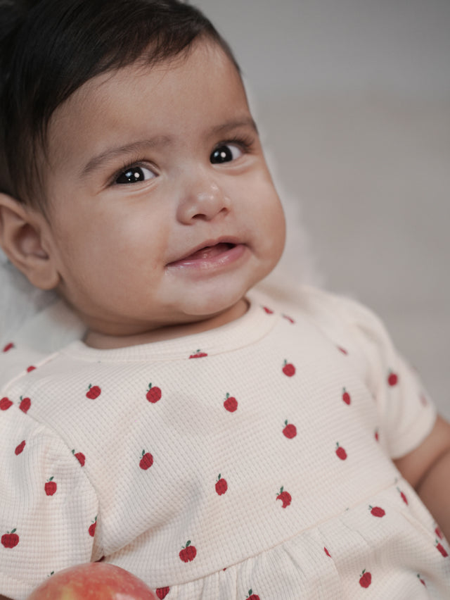 Baby wearing cream organic cotton dress with red apple print, GOTS certified baby clothing by Mamma & Amour
